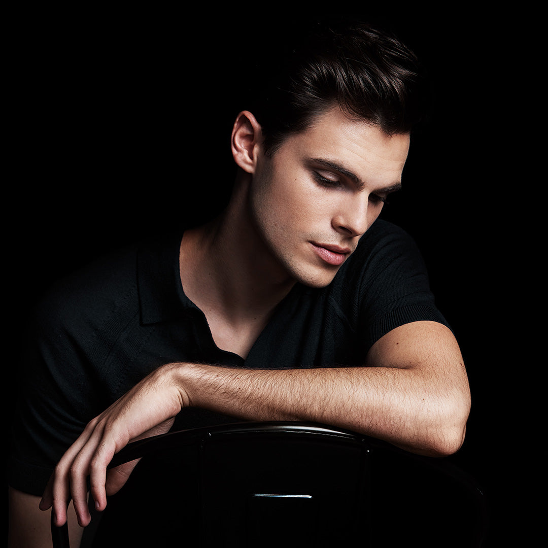 Image of male model sitting and resting his arm over a chair looking down and showing off his bold brows 