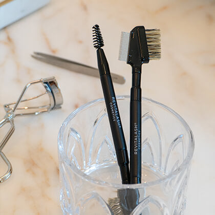 Two black eyebrow brushes with bristle heads in a clear glass holder on a marble surface.
